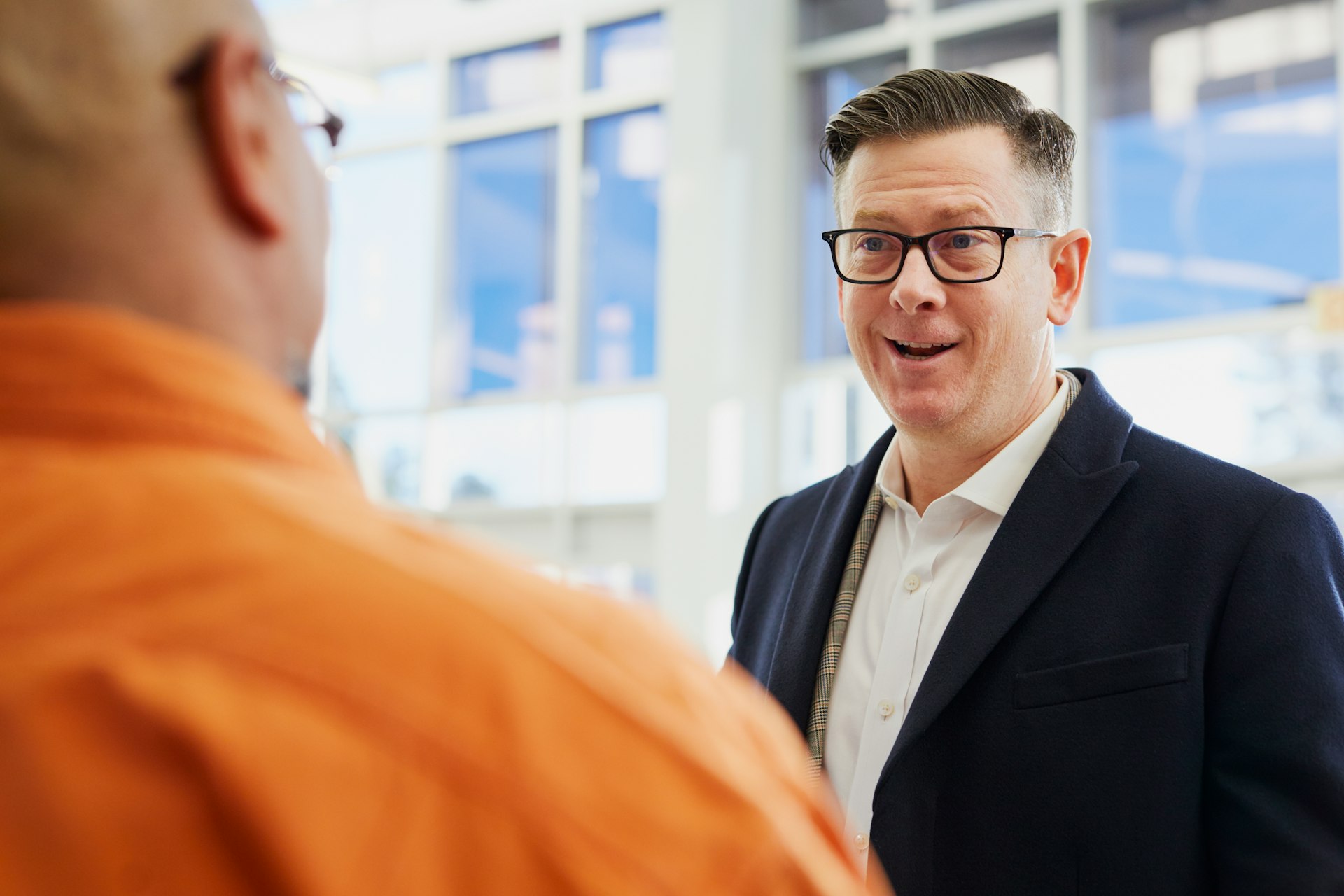 Conversation Professionnelle dans un Cadre d'Affaires Un homme habillé professionnellement avec des lunettes échange dans une conversation amicale avec une autre personne en milieu professionnel.